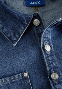 Zbliżenie niebieskiej dżinsowej kurtki z metalowymi napami JJXX oraz pasującą niebieską metką JJXX wewnątrz kołnierza.