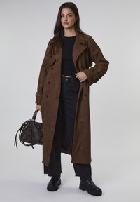 Kobieta w długim brązowym płaszczu z zamszu, czarnym, krótkim topie, czarnych dżinsach i masywnych czarnych botkach, trzymająca czarną skórzaną torebkę, stojąca na jednolitym tle.