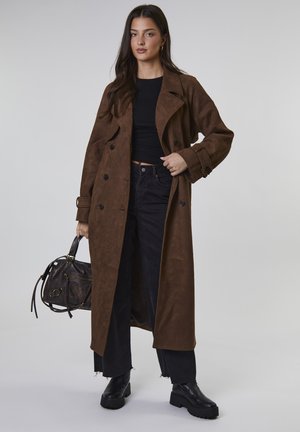 Frau mit einem langen braunen Wildledermantel, schwarzem Cropped-Top, schwarzen Jeans und schwarzen klobigen Stiefeln, die eine schwarze Ledertasche hält und vor einem einfarbigen Hintergrund steht.