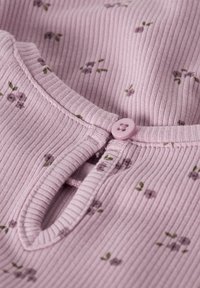 Gros plan sur un tissu rose côtelé avec de petits motifs floraux violets et un bouton rose assorti au-dessus d'une ouverture en forme de goutte d'eau au niveau du décolleté.