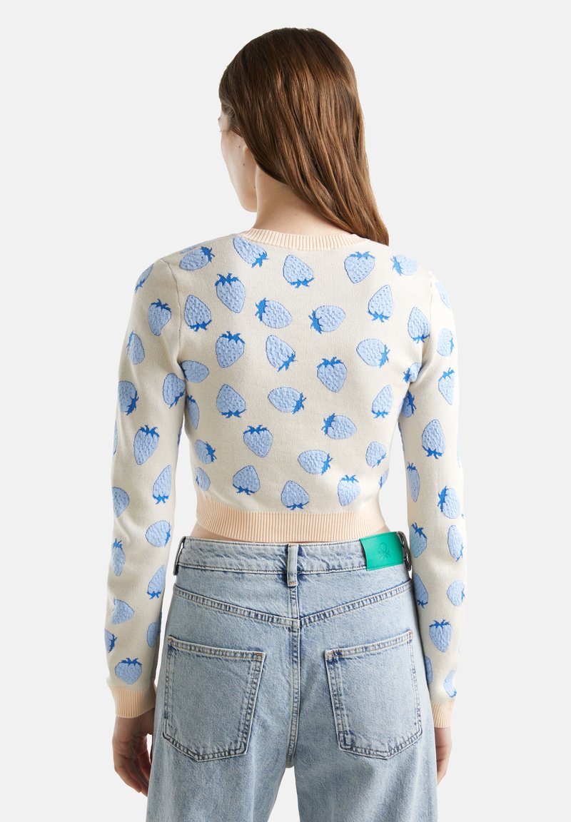 Kremowy sweter z jasNONB niebieskim wzorem truskawek i ściągaczami, zestawiony z jasnoniebieskimi dżinsami z widoczną zieloną metką na pasie.