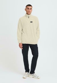 Fleece-Pullover in hellem Creme mit einem hohen Kragen, ausgestattet mit einem Reißverschluss und einem Patch-Label. Kombiniert mit schwarzen Cargo-Hosen und weißen Sneakers.