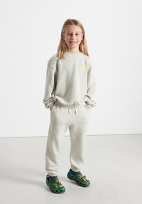 Šedá flísová mikina s malým žltým logom, skombinovaná s ladiacimi zúženými teplákmi. Má na sebe zelené kamuflážne šľapky s modrými akcentmi.