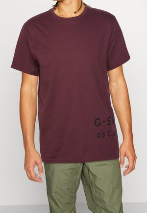 T-shirt en coton marron avec un col rond, des manches courtes et un texte imprimé noir en bas à gauche. Porté avec un pantalon vert olive.