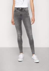 Person i urtvättade grå skinny jeans, vita sneakers och en vit ärmlös topp står mot en enfärgad bakgrund.