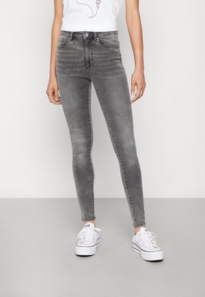 Persoon in vervaagde grijze skinny jeans, witte sneakers en een witte mouwloze top, staand tegen een effen achtergrond.