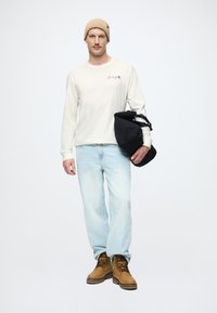 Camisa de manga larga beige claro con logo, jeans azul claro, botas marrones y un gorro beige. Sosteniendo una bolsa negra. Fondo blanco liso.