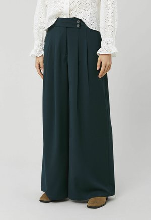 Femme portant un pantalon large vert foncé avec des plis à l'avant, une taille à double bouton, associé à un chemisier blanc à œillets et des chaussures marron.