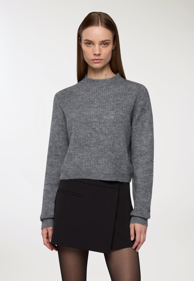 Kobieta z prostymi brązowymi włosami, nosząca szary sweter z dzianiny w połączeniu z czarną asymetryczną mini spódnicą i czarnymi rajstopami.