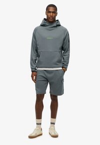 Sudadera gris con cuello alto, bolsillo frontal y logo verde en contraste. Combinada con pantalones cortos a juego, que presentan un bolsillo lateral y un diseño deportivo.