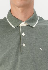 Groene polo met een gestructureerde stof, voorzien van een contrasterende lichtgekleurde kraag, drie knopen en een klein logo op de borst.