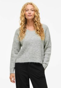 Pull gris tricoté avec un décolleté en V, des manches longues et un ourlet légèrement cropped ; texture douce avec des détails de tricot lâche. Associé à un pantalon noir.