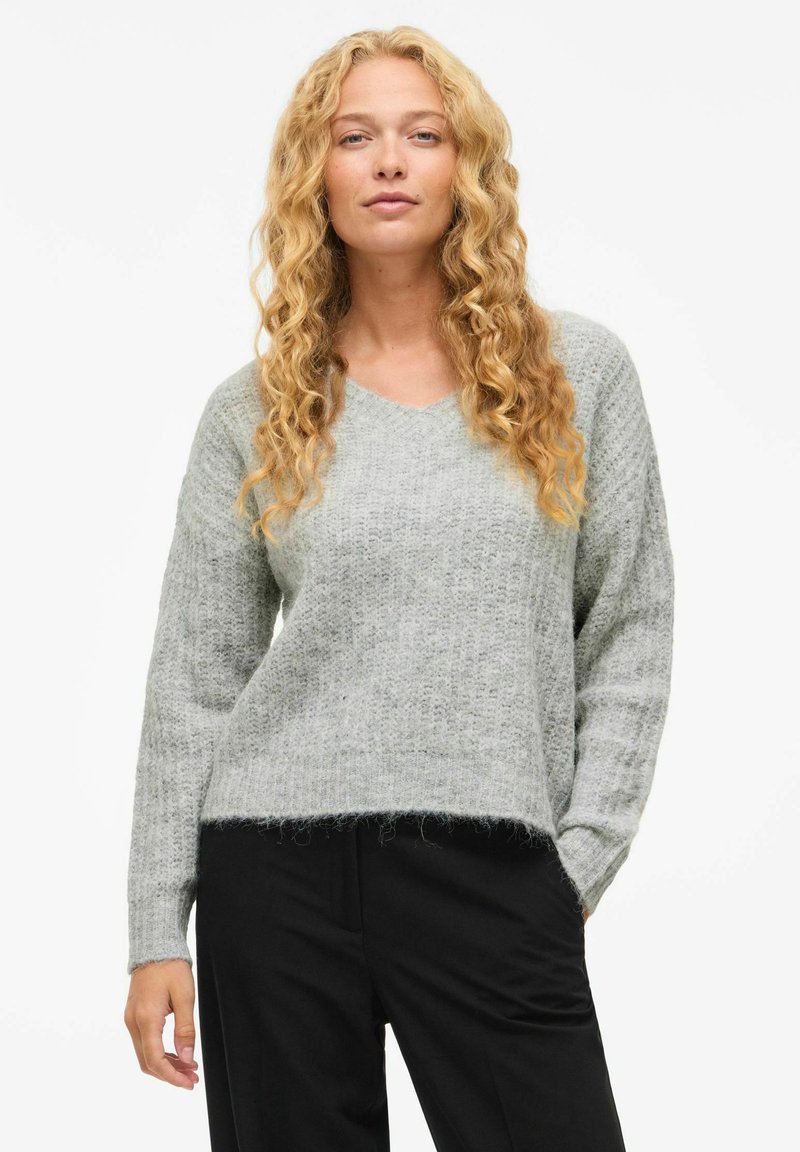 Pull gris tricoté avec un décolleté en V, des manches longues et un ourlet légèrement cropped ; texture douce avec des détails de tricot lâche. Associé à un pantalon noir.