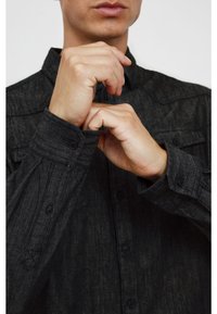 Tummanharmaa nappikauluspaita, jossa on teksturoitu kangas, esiteltynä niin, että kädet säätävät hihansuita, ja siinä on terävä kaulus sekä taitetut hihansuut.