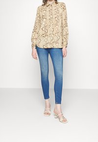 Blusa de estampado de serpiente beige con cuello, mangas largas y botones, combinada con jeans ajustados azules y sandalias de tacón blancas con tiras.