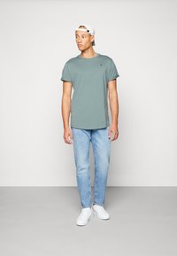 Camiseta verde de corte relajado y manga corta con un dobladillo redondeado y un pequeño logo. Combinada con vaqueros de color azul claro y zapatillas blancas.