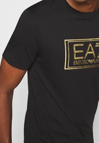 Svart bomullströja med guldfolie-logotypdesign. Klassisk rundhals med korta ärmar, slät textur och figurnära passform.