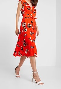 Robe mi-longue rouge à fleurs avec un col à volants, une taille cintrée et une jupe évasée. Associée à des escarpins blancs à bride à la cheville.