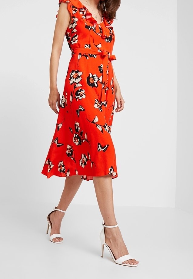 Robe mi-longue rouge à fleurs avec un col à volants, une taille cintrée et une jupe évasée. Associée à des escarpins blancs à bride à la cheville.