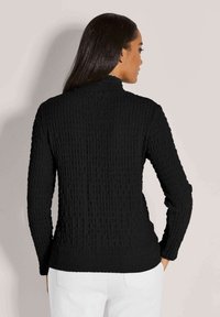 Frau mit langen dunklen Haaren, die ein schwarzes, strukturiertes Langarmoberteil und weiße Hosen trägt, mit dem Rücken zu einem schlichten, hellen Hintergrund.