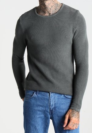 Uomo che indossa un maglione a maniche lunghe di colore grigio scuro testurizzato e jeans blu, con tatuaggi visibili sul collo e sulle mani, in piedi davanti a uno sfondo semplice.