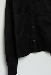 Giacca in suede nera con bottoni a pressione, due tasche sul petto e orlo leggermente accorciato. Tessuto morbido, design minimalista.