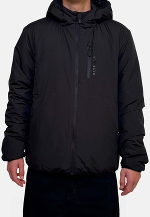 Personne portant une doudoune noire à capuche avec une fermeture éclair frontale et une poche poitrine verticale zippée portant l'étiquette "REDSKINS."