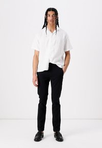 Chemise blanche en lin à manches courtes, pantalon noir slim, et mocassins noirs. Le mannequin se tient sur un fond neutre, les mains dans les poches.