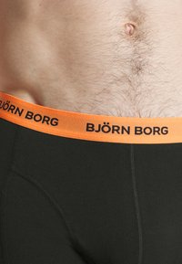 Čierne spodné prádlo s jasne oranžovým elastickým pásikom, na ktorom je potlač "BJÖRN BORG" v čiernej farbe. Tkanina vyzerá hladko a priliehavo.