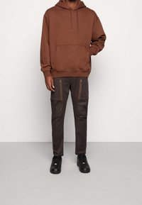 Ruskea huppari, jossa on kengurutasku, yhdistettynä tummanruskeisiin cargo-housuihin, joissa on vetoketjulliset taskut ja sileä pinta. Mustat lenkkarit.