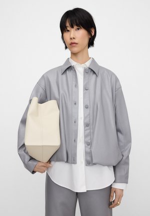 Kobieta o krótkich czarnych włosach nosi szarą kurtkę zapinaną na guziki na białą koszulę i szare spodnie, trzyma dużą beżową torbę na ramię.