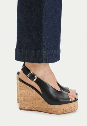 Füße, die schwarze Leder-Wedges mit offenen Zehen, Korkabsätzen und goldenen Schnallenriemen tragen, kombiniert mit dunkelblauen Jeanshosen.