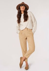 Creme kabelstrik sweater med ribbede manchetter; parret med beige bukser med høj talje og to-knaps lukning; sat mod en neutral baggrund.
