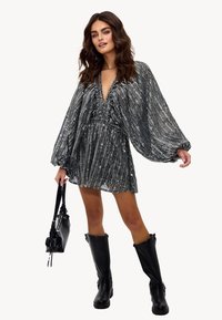 Robe mini rayée noire et argentée avec des manches amples, décolleté en V profond et texture plissée. Associée à des bottes noires jusqu'aux genoux et un sac à main.