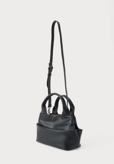 Μαύρη δερμάτινη τσάντα χειρός με δύο κοντές λαβές, μακρύ ρυθμιζόμενο λουράκι ώμου και μεγάλη μπροστινή τσέπη, όρθια σε λευκό φόντο.