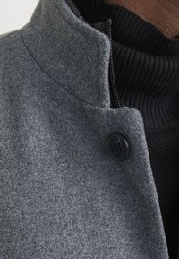 Gros plan sur un manteau en laine gris avec un bouton noir sur un pull côtelé noir et une peau marron visible.