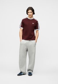 Νεαρός άνδρας που στέκεται με τα χέρια στις τσέπες, φοράει ένα μπορντό μπλουζάκι Adidas με λευκές ρίγες, γκρι φόρμα και μπλε αθλητικά παπούτσια.