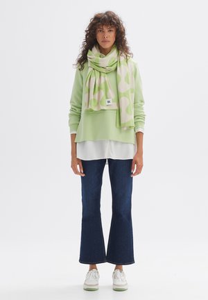 Hellgrüner, übergroßer Pullover über einer weißen Bluse, kombiniert mit dunkelblauen, ausgestellten Jeans und einem gemusterten Schal in Grün und Beige.