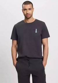 Czarnoszara bawełniana koszulka z krótkim rękawem, okrągłym dekoltem i jasnoniebieskim elementem graficznym na klatce piersiowej. W zestawie z ciemnymi dresami.