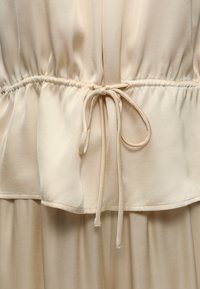 Ενδύματα από μπεζ ύφασμα με δεμένο κορδόνι στη μέση, ραφές με πτυχώσεις και απαλό, ρέον ύφος.