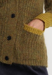 Käsi kollaste puffidega taskus paksu pruun-roheline kudumiskardigan, milles on mustad nööbid, seljas tumedate ribakudumistega pükste peal.