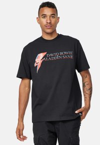 Svart bomull t-shirt med texten "David Bowie Aladdin Sane" och en blixtgrafik i röd och blå. Loose fit med korta ärmar.