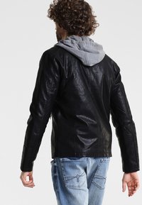 Veste en cuir noir avec doublure de sweat à capuche grise, présentant une coupe ajustée et des épaules structurées, vue de dos avec un jean en denim.
