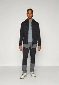 Crna pletiva jakna s patentnim zatvaračem, sivi dugi rukav i siva hlače s kontrastnim crnim flasterima. Bijele tenisice upotpunjuju odjevnu kombinaciju.