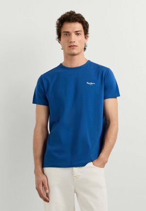 Jeune homme aux cheveux bouclés portant un t-shirt bleu à manches courtes et un pantalon clair, debout contre un mur clair uni.