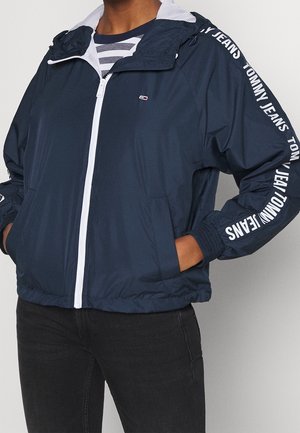 Veste bleu marine à fermeture éclair blanche, texte "Tommy Jeans" sur les manches, portée par-dessus une chemise rayée, mains dans les poches.