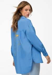 Light blue oversized shirt made of smooth fabric with long sleeves, featuring yellow embroidered designs on the back and side slits.
