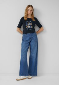 T-shirt graphique marine à manches courtes, arborant l'inscription "BONNES CHOSES À VENIR" en lettres majuscules, associé à un jean bleu à jambes larges et des baskets blanches.