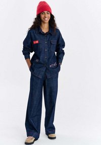 Kurtka dżinsowa i spodnie o szerokich nogawkach w ciemnym niebieskim kolorze, z czerwonym przeszyciem i dwoma przednimi kieszeniami ozdobionymi zabawnymi wzorami. Modelka ma na sobie czerwoną czapkę typu beanie.