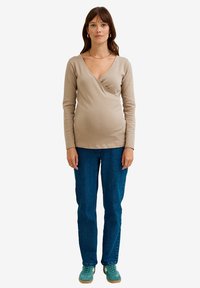 Beige langärmliges Wickeloberteil mit V-Ausschnitt, kombiniert mit blauen Jeans und türkisfarbenen Sneakern. Einfaches Design, weiche Textur und keine sichtbaren Muster oder Akzente.
